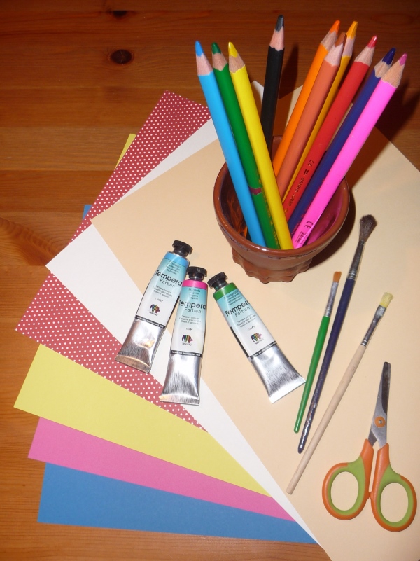 Auf dem Tisch liegen Pinsel, Farben, Schere und buntes Papier zum Basteln.