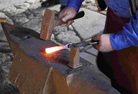Ein Mann hält mit der Zange ein glühendes Metallstück in Position, gleich folgt ein Schlag mit dem Hammer.