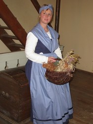 Eine bäuerlich gekleidete Frau hält einen dunkelbraunen Korb in der Hand. Er ist gefüllt mit einem Wildblumenstrauß.