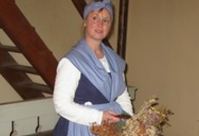 Eine bäuerlich gekleidete Frau hält einen dunkelbraunen Korb in der Hand. Er ist gefüllt mit einem Wildblumenstrauß.